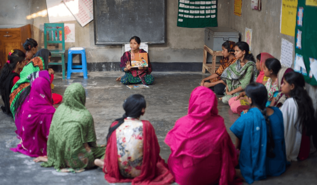 A woman with light brown skin and black hair in green dress and red and black shawl sits at the front of a classroom teaching 16 young women who are seated around her in a horseshoe shape