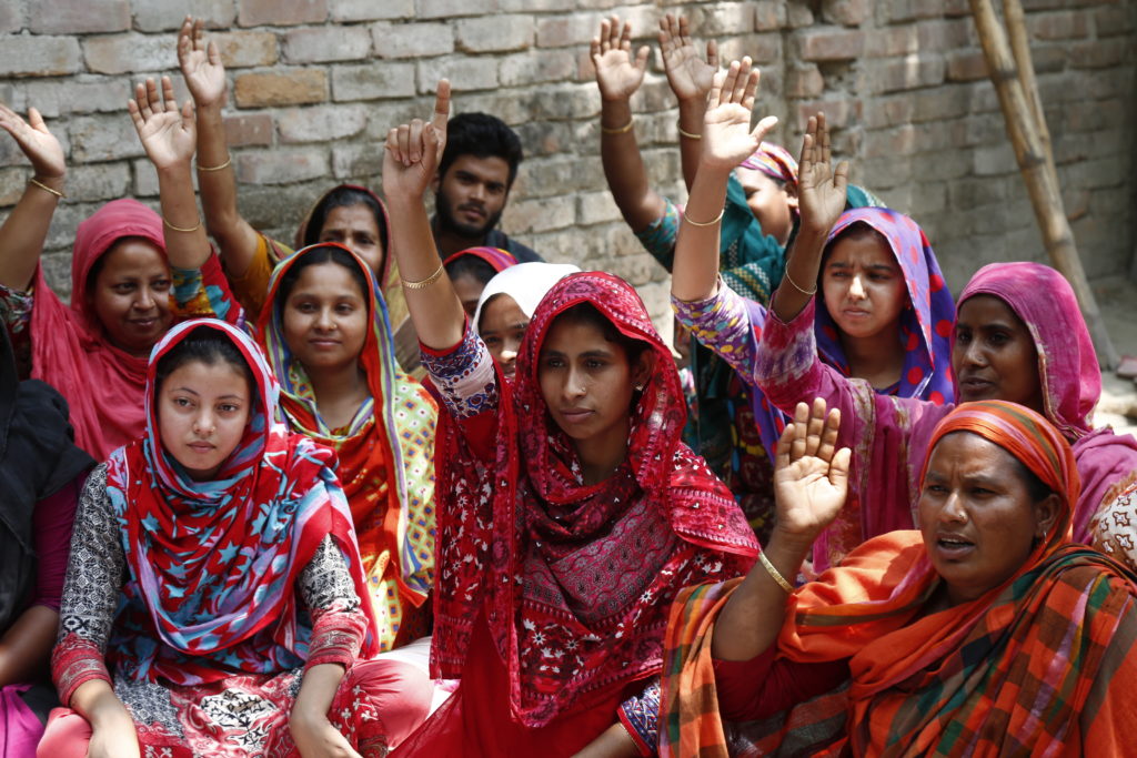 A group of Brown women wearing colourful saris are sitting down on the floor and raising their hands.