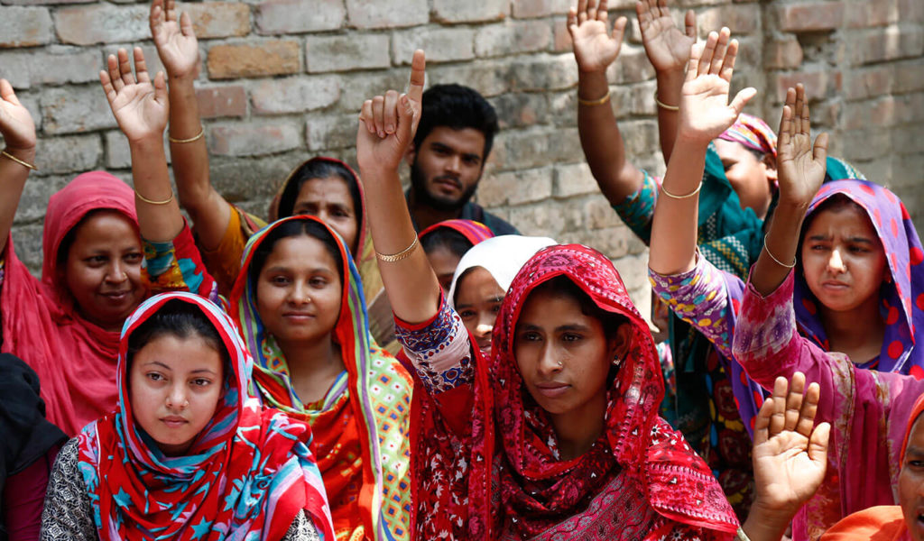 A dozen young brown-skinned women are seated together outdoors in the daytime. They are all wearing multi-coloured head scarves and brightly-coloured dresses. Most of them have their hands up as if they are all waiting to be called on by a teacher.