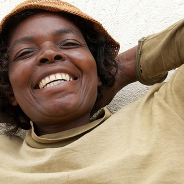 A dark-skinned, smiling woman leans back with her hands behind her head. She wears a long-sleeved, beige shirt and a brown bucket hat.