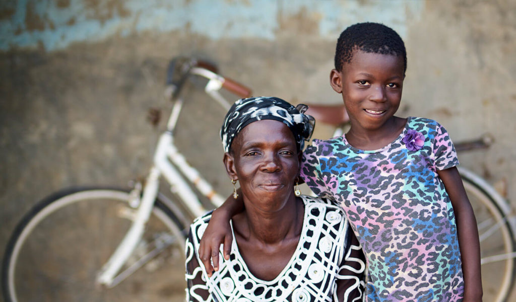 A dark-skinned older adult in a black-and-white dress and a head scarf is seated on a porch, and a young, dark-skinned feminine-presenting child stands next to her with her arm around the adult. They are both smiling. The child wears a pastel-coloured animal print t-shirt. Behind them, a white bicycle leans against a damaged concrete wall.