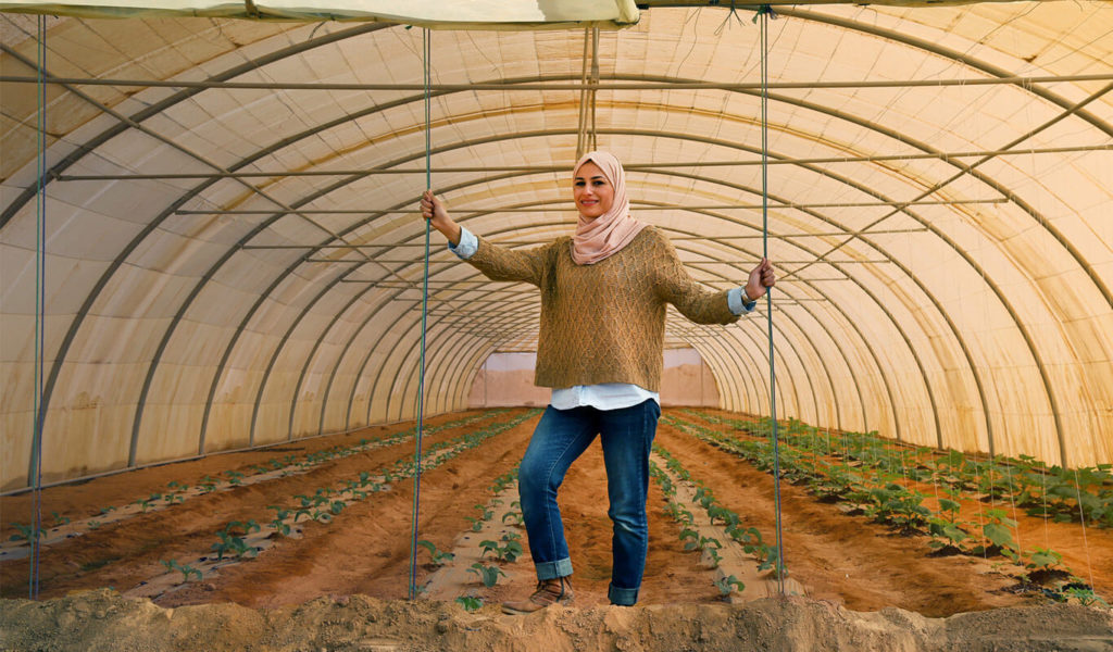 A young woman is standing inside a greenhouse between two rows of vegetables that are growing out of the ground under the cover of a white dome. She is smiling and wearing a pink head scarf, a white long-sleeved t-shirt with a light brown sweater over it, a pair of dark blue jeans that are rolled up at the bottom and brown boots. She is holding onto two ropes that are hanging down from the top of the greenhouse structure.