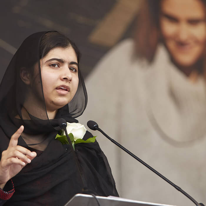 Nobel Peace Prize winner Malala is shown speaking at a public event. She is dressed in a black dress and black head scarf and is standing in front of a microphone that is attached to a podium. Behind her are pictures of women.