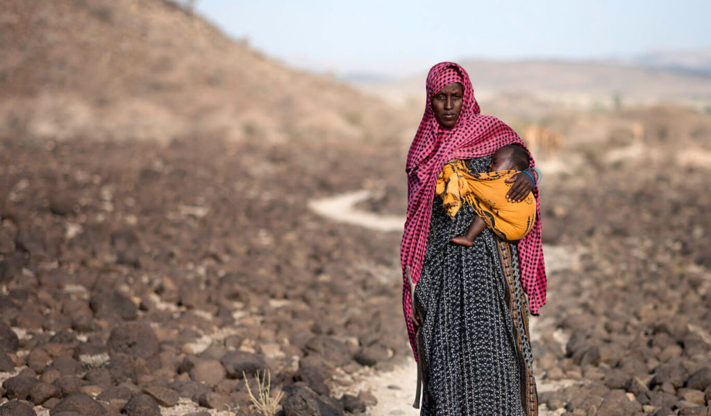 A dark-skinned young woman in a long, black-and-white patterned dress and a red and black head scarf is standing in a drought-ridden field while her baby sleeps in a yellow and black sling.