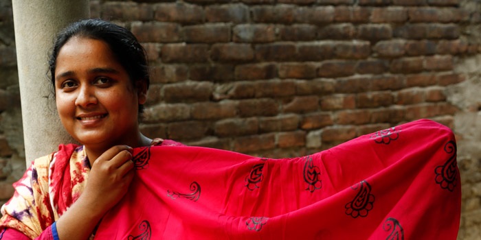 A young brown-skinned woman holds up red fabric with a dark blue pattern on it. She is outdoors and smiling, and it's daytime.