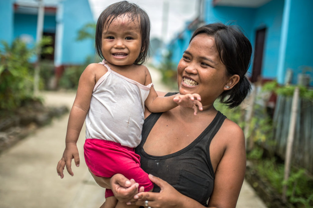 A smiling woman carries her laughing toddler in her arms while standing on a street surrounded by tropical plants and blue houses.