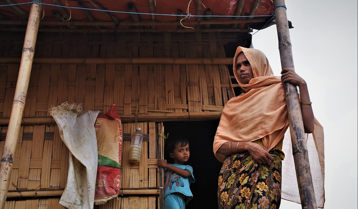 Women in an orange headscarf and her sun standing in front of their shelter in Bangladesh