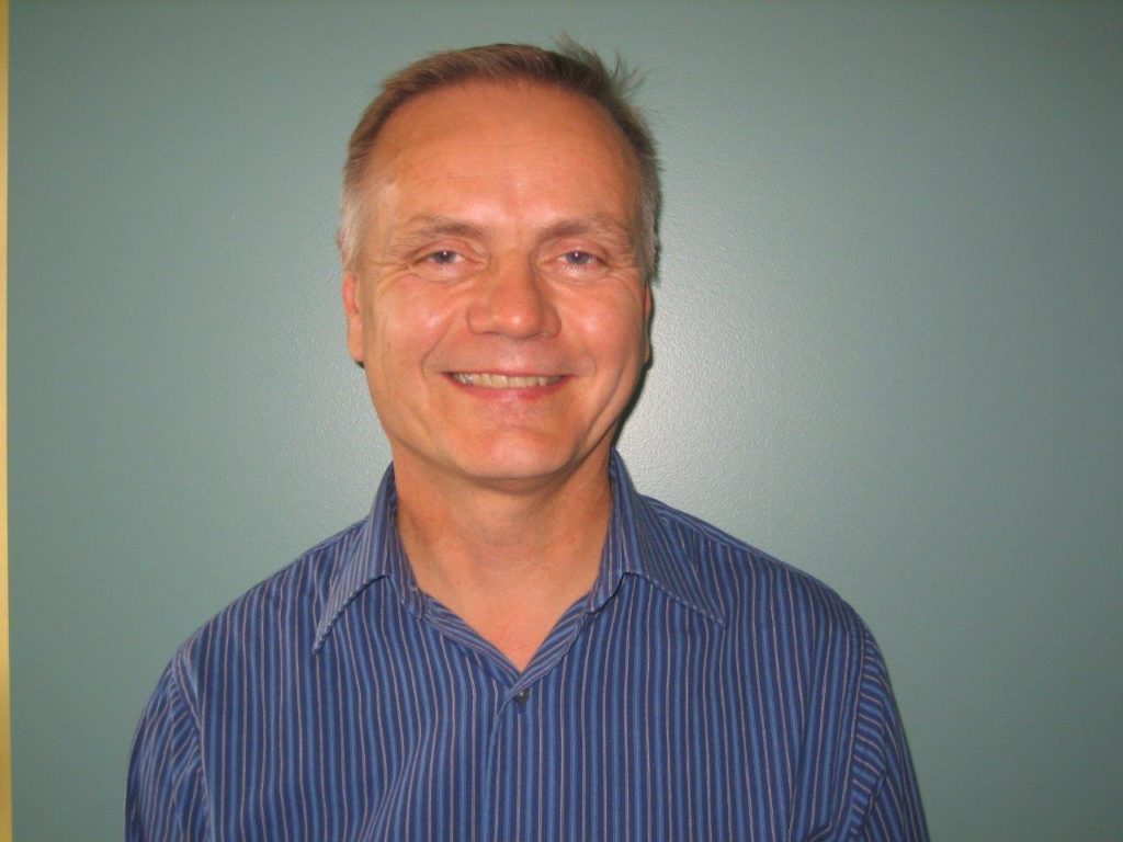 A headshot of Kent MacAulay smiling. A white man in a blue shirt and grey hair against a grey background.