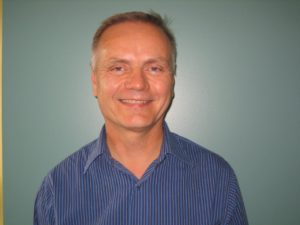 A headshot of Kent MacAulay smiling. A white man in a blue shirt and grey hair against a grey background.