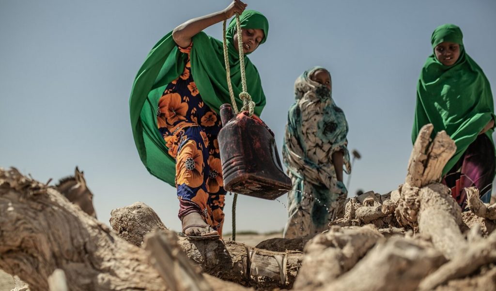 Three young girls dressed in green hijabs and colourful dresses standing around a hole in the dry, hard ground with dead logs around it. One girl is holding a black watering can with a rope tied to it and it has water leaking from the bottom.eces of 