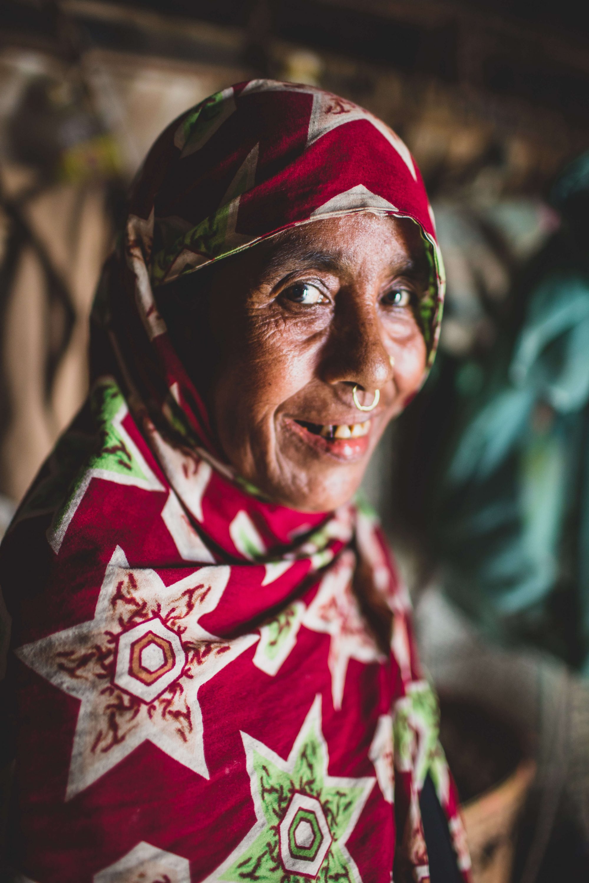 Noon_Nahar_Rohingya Portrait of a woman wearing a colourful red and green veil and a ring nose, looking directly at the camera, smiling.