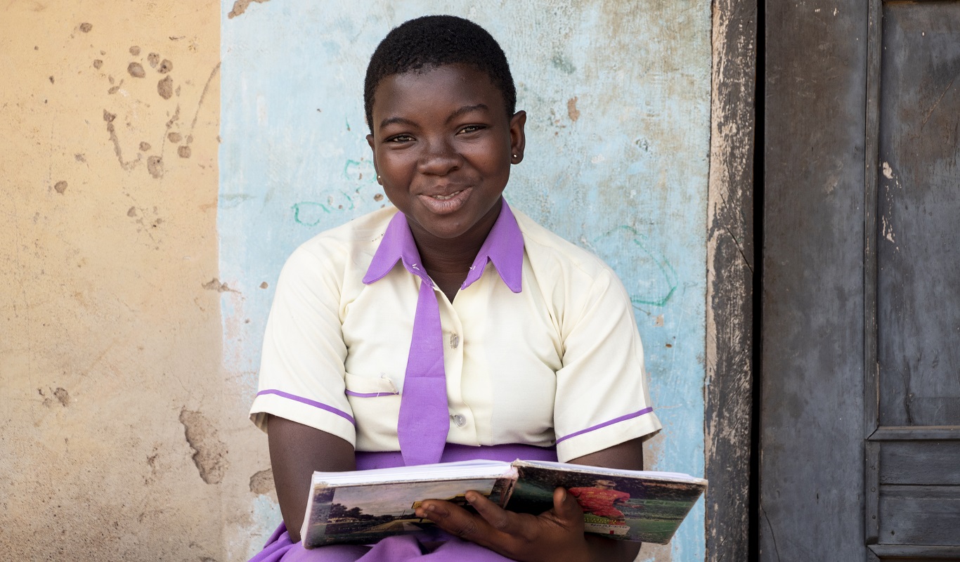 Haddisah Ibrahim studying - Ghana - 16894 A young school girl in a white and purple school uniform smiles with a book open on her lap.