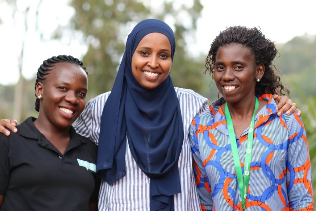 Three dark-skinned women stand together. The woman in the middle is wearing a blue head scarf and has her arms around the other two women. They are all outside during the daytime and all are smiling. The woman on the left is wearing a black Oxfam T-shirt and the woman on the right is wearing a green and white Oxfam lanyard. They are all Oxfam staff.