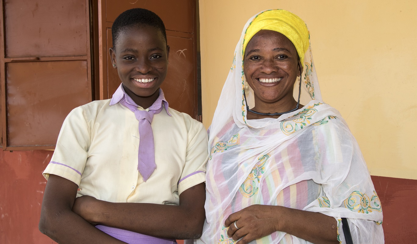 A school girl in a purple and white school uniform stands next to her mother. They are both smiling.