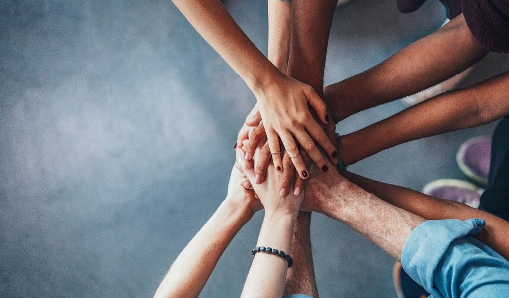 Nine hands of different races and ages are piled on top of each other.