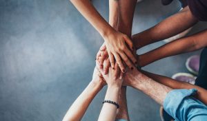 Nine hands of different races and ages are piled on top of each other.
