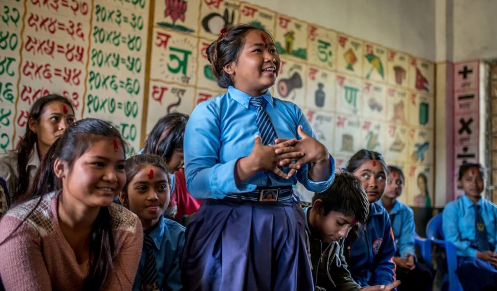 One young girl in a school uniform is smiling and laughing as she stands up amongst a group of other children in a classroom