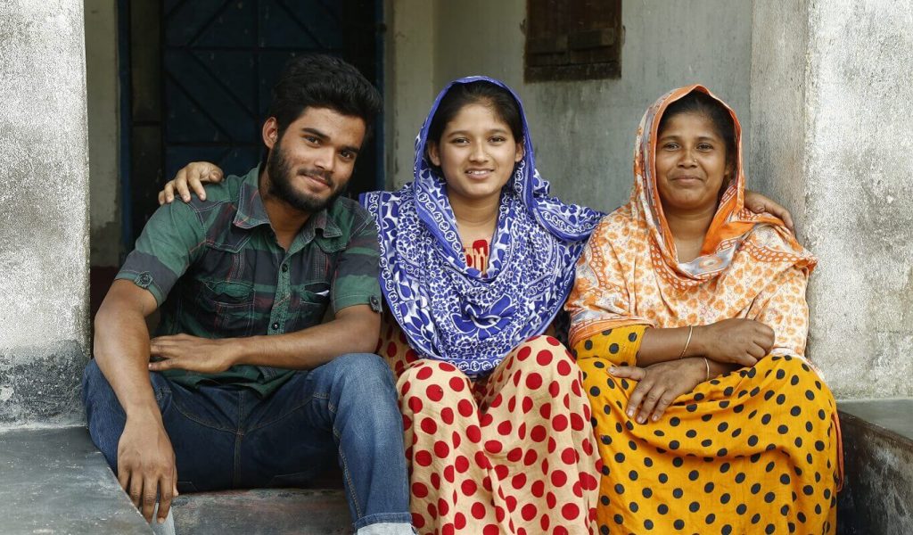 A young man, a young woman and their mother sit on the front step of their home with their arms around each other.