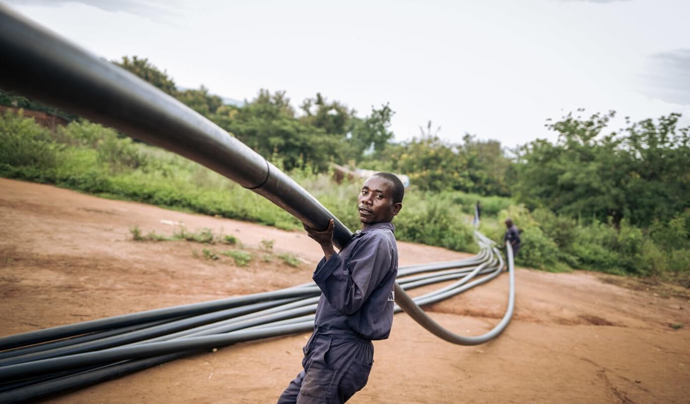A black man in coveralls covers a long, thick water pipe. On the ground, there are several more pipes.
