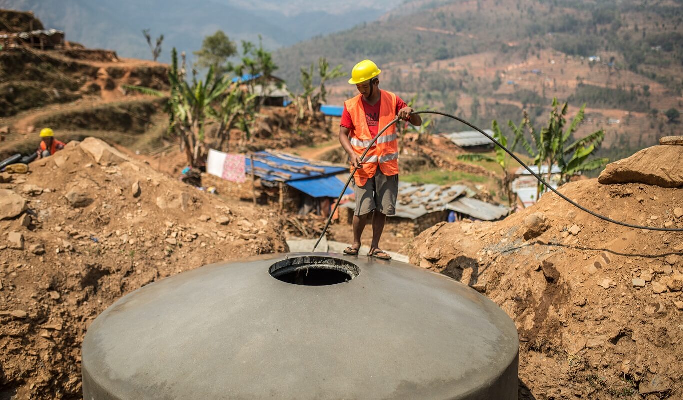 A man in a yellow hardhat and construction vest feeds a wire down into a large water supply container sitting in a hole in the ground in Nepal.