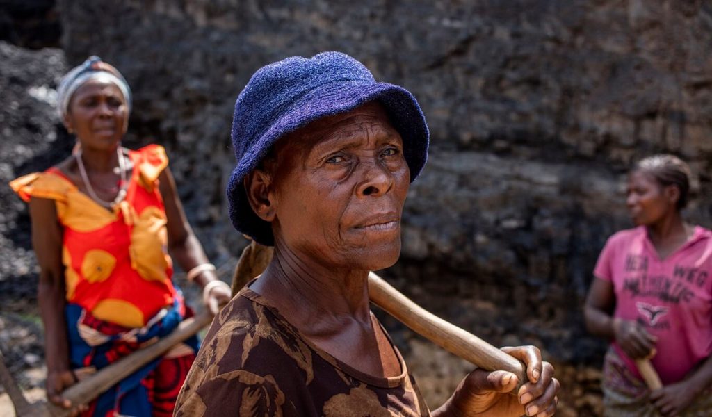 An elderly black woman in a blue hat stands with a pickaxe over her shoulder and a serious look on her face.