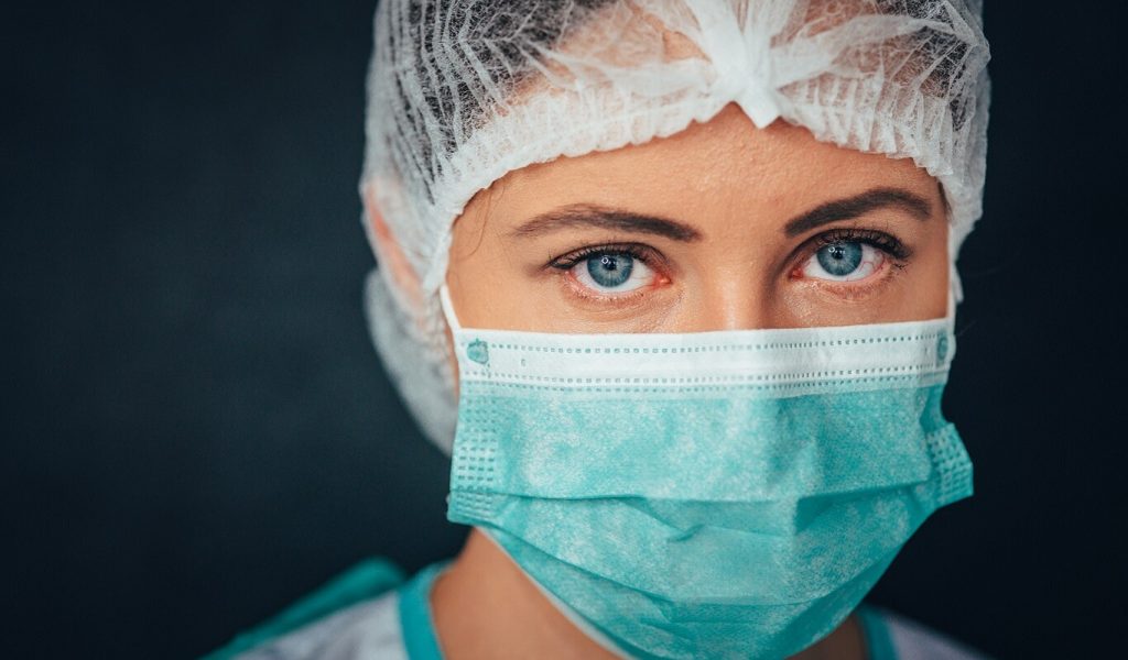 A health care worker in a protective mask and hair cover and scrubs and gown looks at the camera.