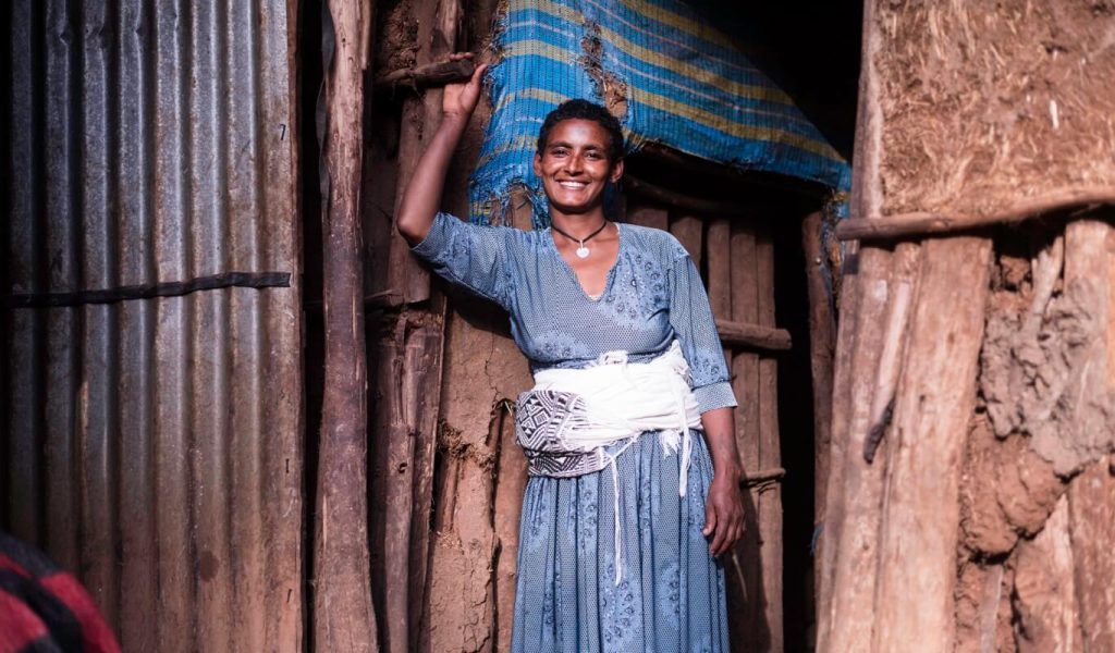 A 30-year-old Ethiopian woman with very short hair in a long blue dress stands smiling in the doorway of her home.