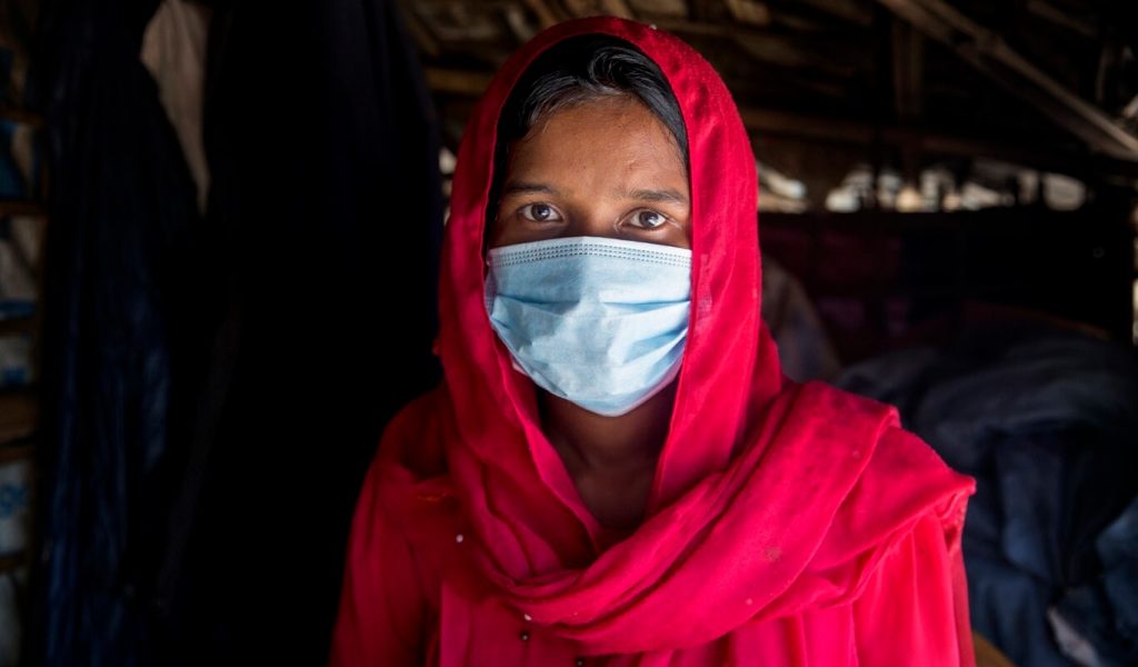 A bangladeshi woman wearing a red head scarf, an N95 mask and a red outfit looks directly at the camera.