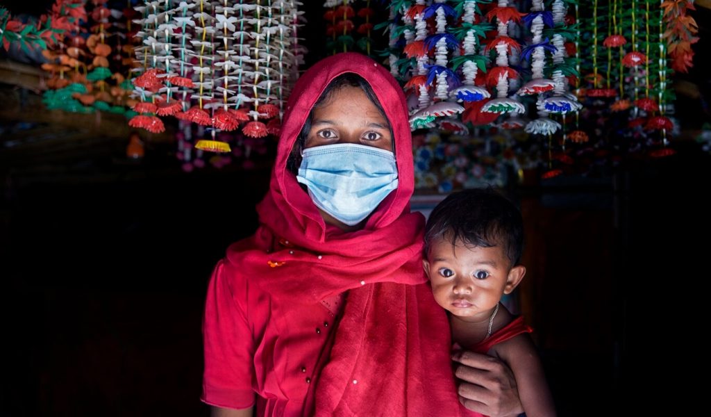 A woman with brown skin, brown eyes and dark hair is holding her young child and wearing a blue surgical mask and a red dress and headscarf.