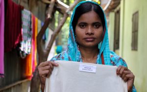 A young woman standing outside in what seems to be a street, holds a white garmen which label rads "Made in Bangladesh." The woman has a solemn expression and is wearing a bright blue headscarf.