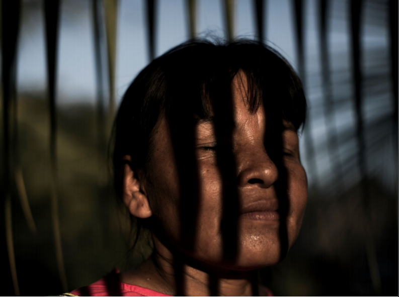 An Indigenous woman with dark hair, pulled back from her face, has closed eyes and lines of vertical shadow across her face