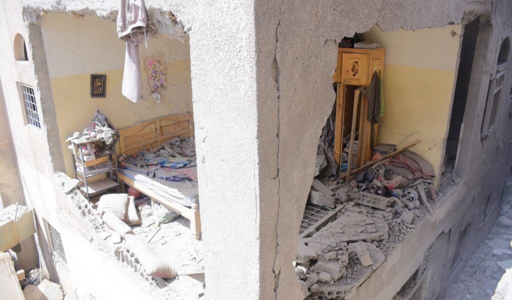 The image shows the inside of a bedroom in San'a that was destroyed by bombing. There is rubble all over the bed, and the external walls have crumbled away.