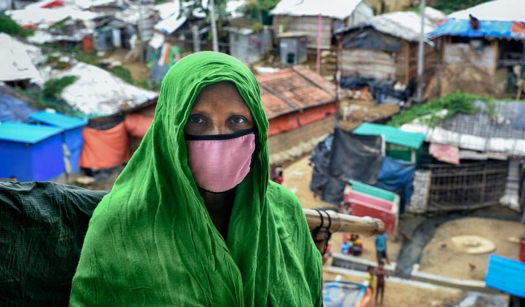 A refugee woman wears a green head covering and a pink cotton mask as she stands in front of a refugee camp