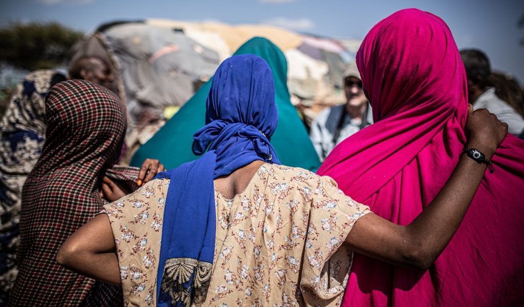 In the foreground of the photo, four brown-skinned women stand with their backs to the camera. Each woman wears a brightly-coloured head scarf. They pose together for a light-skinned, masculine presenting photographer, who can be seen in the background of the photo.