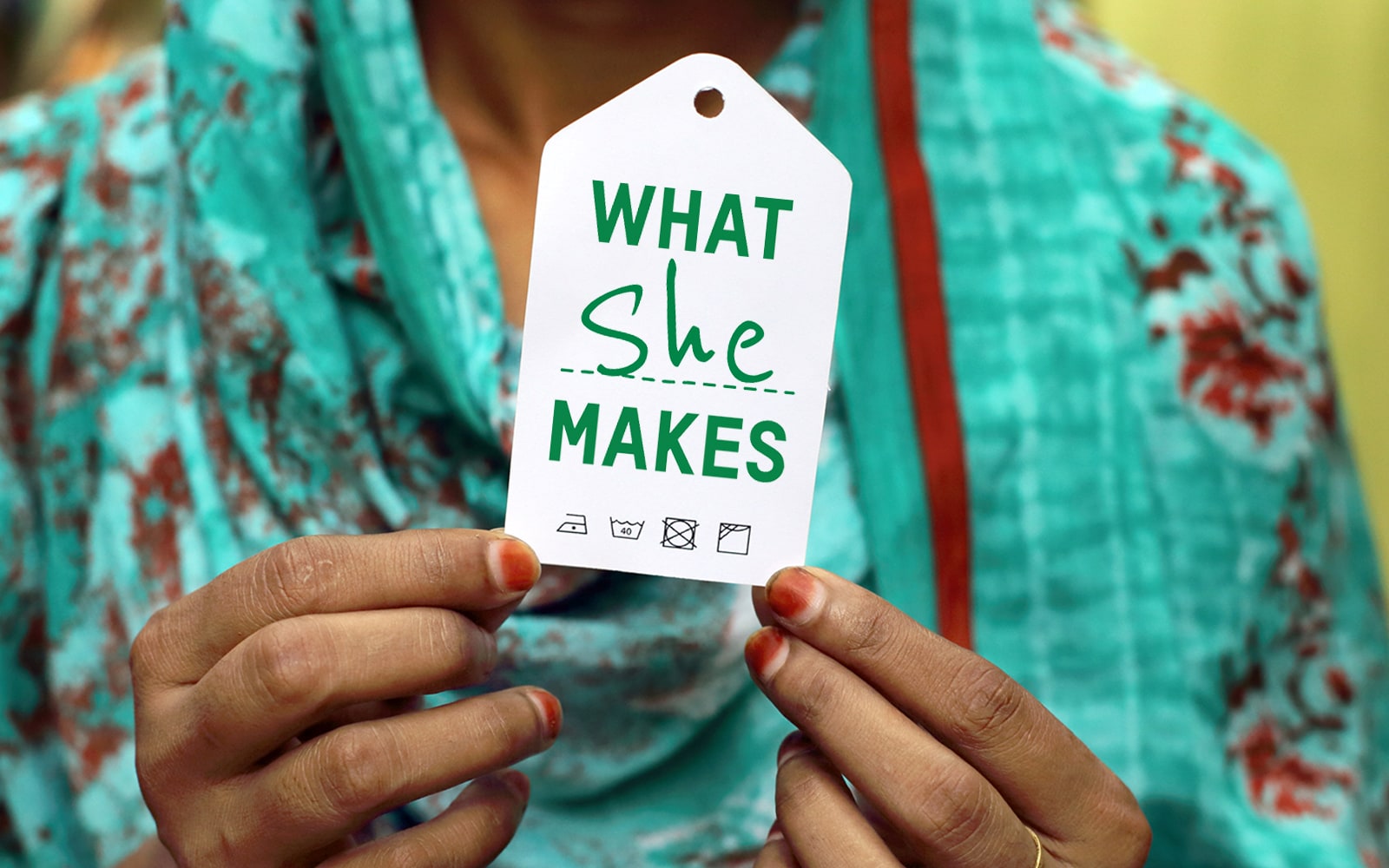 This photo is taken from the neck down. A woman in a teal and red head scarf and red painted nails hold a clothing tag that read