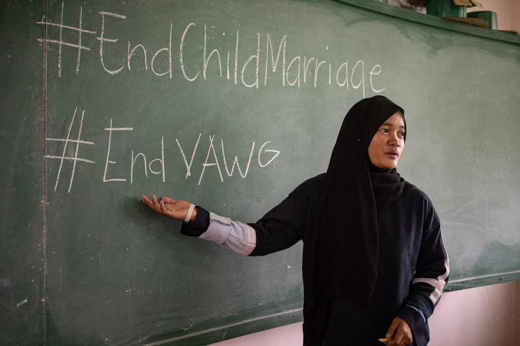 A young Muslim Filipino woman dressed in black stands in front of a chalkboard pointing to the words End Child Marriage and End VAWG