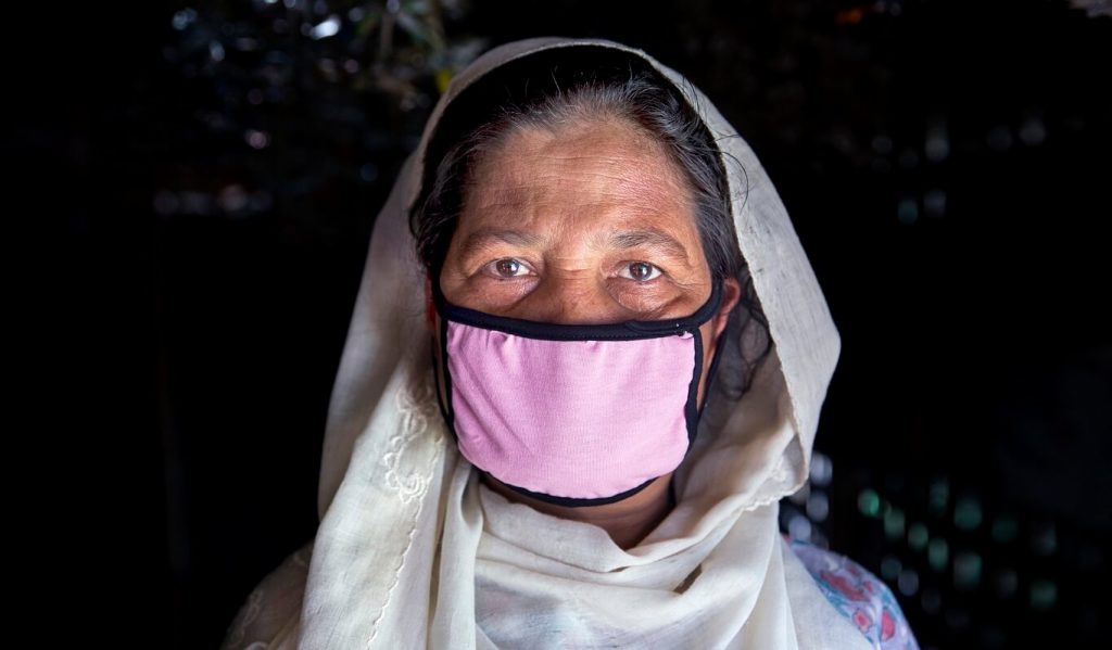 An elderly woman with brown skin, brown eyes and dark hair wears a pink cloth mask with black piping, an off-white head scarf and a floral dress. She is looking directly at the camera.