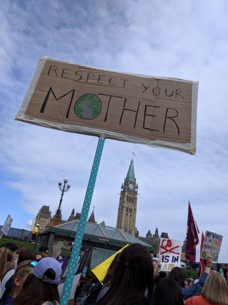 People are gathered in front of the Parliament Buildings in Ottawa and holding signs during climate march. One sign is in the foreground and says Respect Your Mother.