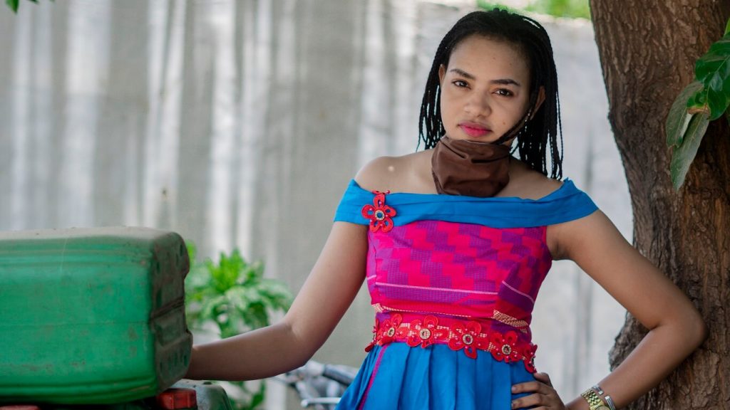 A young Black woman in a multi-coloured off-the-shoulder dress is standing next to a water jug with one hand on her hip and a mask hanging loosely around her neck.