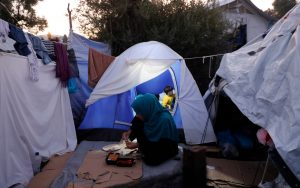 A woman preparing a meal at dusk surrounded by blue and white tents in a refugee camp