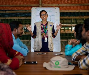 A woman wearing an Oxfam vest and lanyard speaking with a group of people around a table with a chart paper behind her