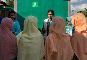 A young woman, speaking to a group of women in front of a green latrine with the Oxfam logo painted on it
