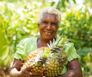 An older woman with white curly hair, smiling and holding three pineapples