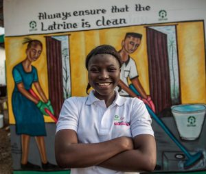 A woman wearing an Oxfam shirt, standing in front of a latrine in Southern Africa