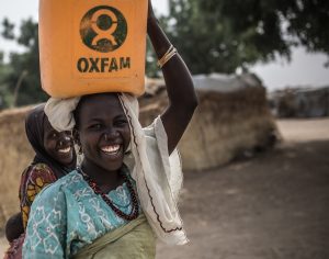 A woman and her friend carrying a yellow jerrycan of water with the Oxfam logo on her head