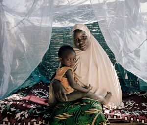 A woman in a pink headscarf holding a child with a mosquito net in the background somewhere in Western Africa