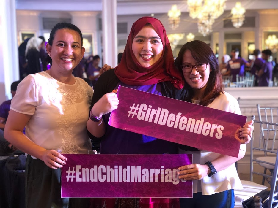 Three girls dressed formally hold two red dark pink banners with white text that reads, hashtag End child marriage and hashtag girl defenders. All the girls are smiling and looking directly at the camera. The one standing in the middle wears a red headscarf.