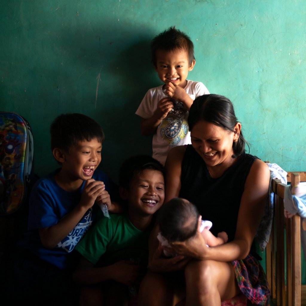 A mother sitting down next to a wooden crib, holds a baby on her lap, smiling a him surrounded by three little boys who are also smiling at the baby. 
