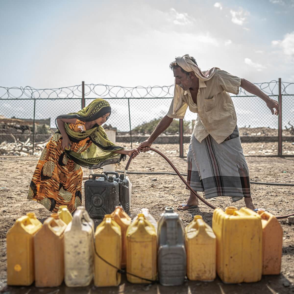 A man and child fill water containers with a hose in front of a barbed wire fence
