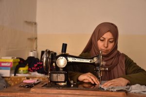 Woman in green dress and mauve hijab sits by her sewing machine.